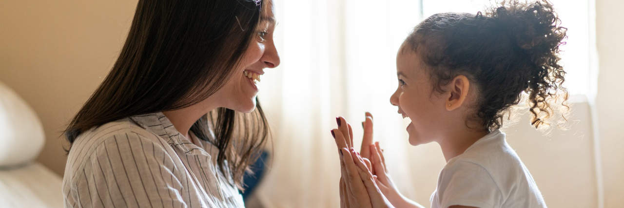 A woman with straight black hair wearing a white striped shirt sits in bed while high-fiving a toddler girl with curly black hair.