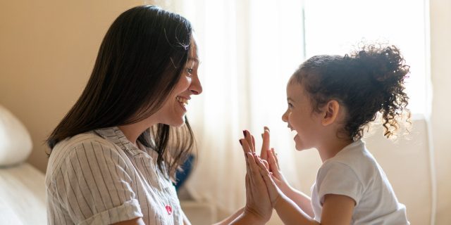 A woman with straight black hair wearing a white striped shirt sits in bed while high-fiving a toddler girl with curly black hair.