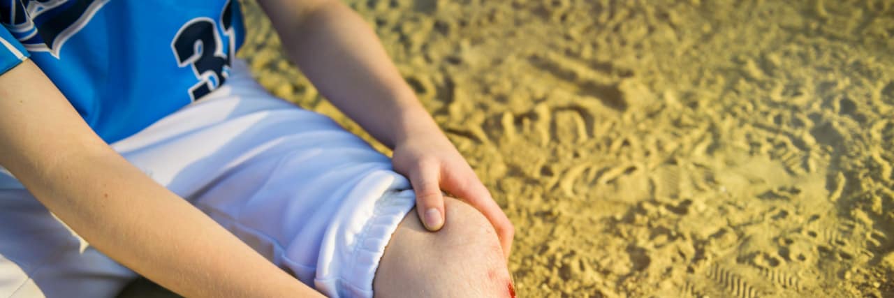 What Playing Little League Baseball Taught Me as a Child With Cerebral Palsy A boy in a baseball uniform sits on a baseball field and has an injury on his leg.