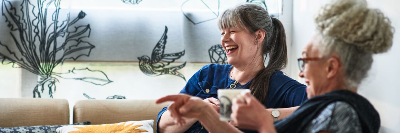 Living Your Best Life With an Alzheimer's Disease Diagnosis Two women sit on a couch and laugh while holding mugs.