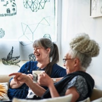 Two women sit on a couch and laugh while holding mugs.