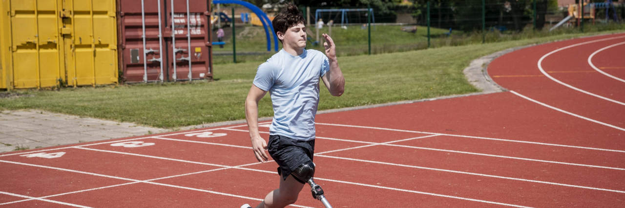 Including Student Athletes With and Without Disabilities in High School Sports A teenage boy with brown hair wearing a light blue shirt, black shorts, and a prosthetic leg runs on a track.
