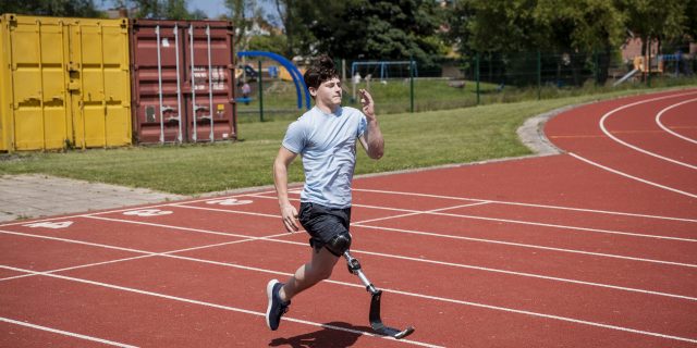 A teenage boy with brown hair wearing a light blue shirt, black shorts, and a prosthetic leg runs on a track.