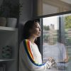A woman with short brown hair wearing a white sweater with multicolored sleeves looks out a window at the rain.
