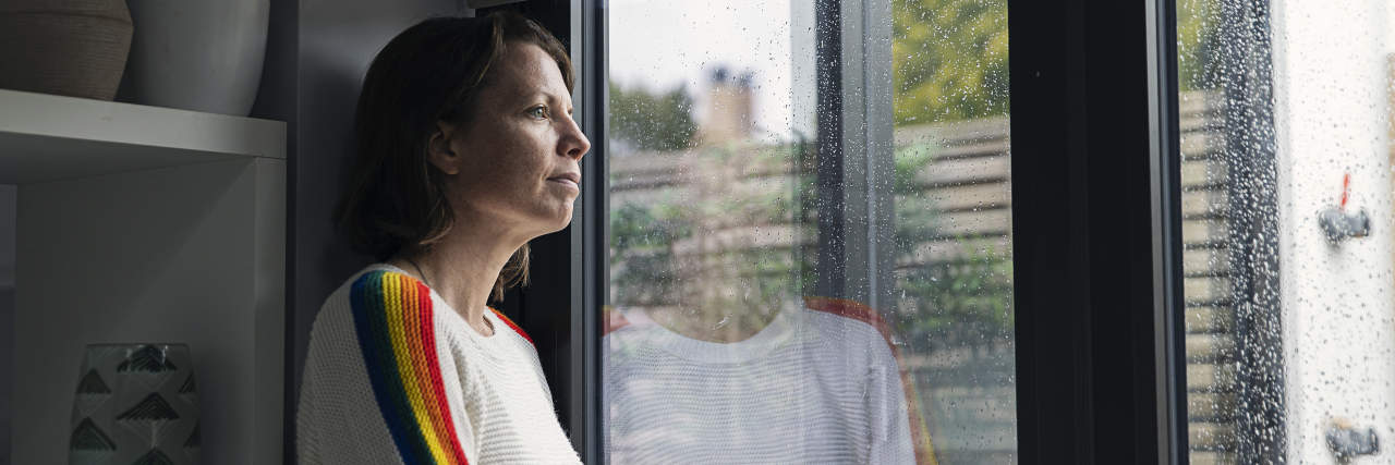 A woman with short brown hair wearing a white sweater with multicolored sleeves looks out a window at the rain.
