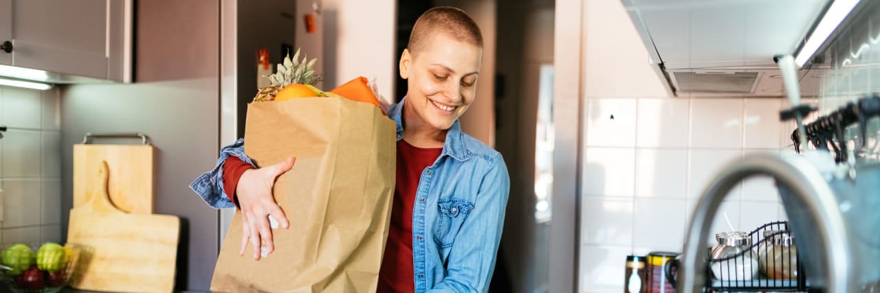 Changing Health Perspectives With Cerebral Palsy and Metastatic Breast Cancer A person with short brown hair stands in a kitchen holding a bag of groceries.