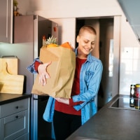 A person with short brown hair stands in a kitchen holding a bag of groceries.