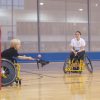 Three people are in an indoor gym. They are all using wheelchairs. They have lacrosse sticks and are playing a match.
