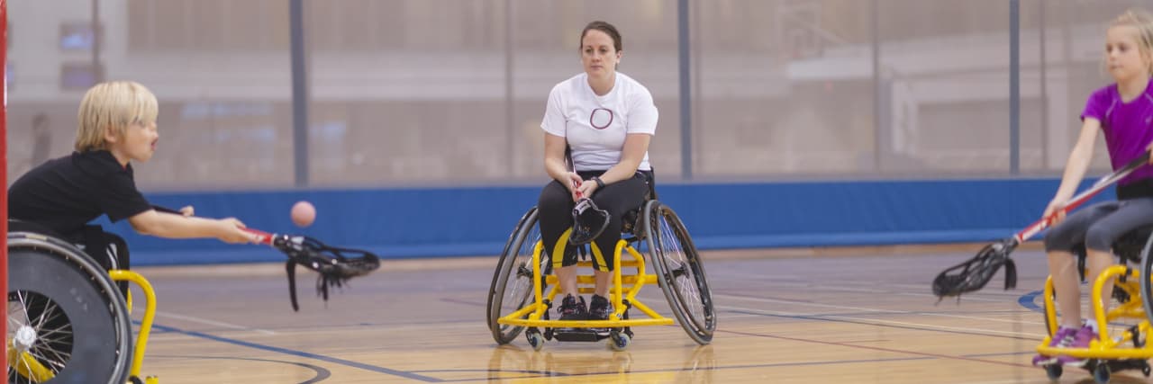 Why Able-Bodied Athletes Should Be Allowed to Play Adaptive Sports Three people are in an indoor gym. They are all using wheelchairs. They have lacrosse sticks and are playing a match.