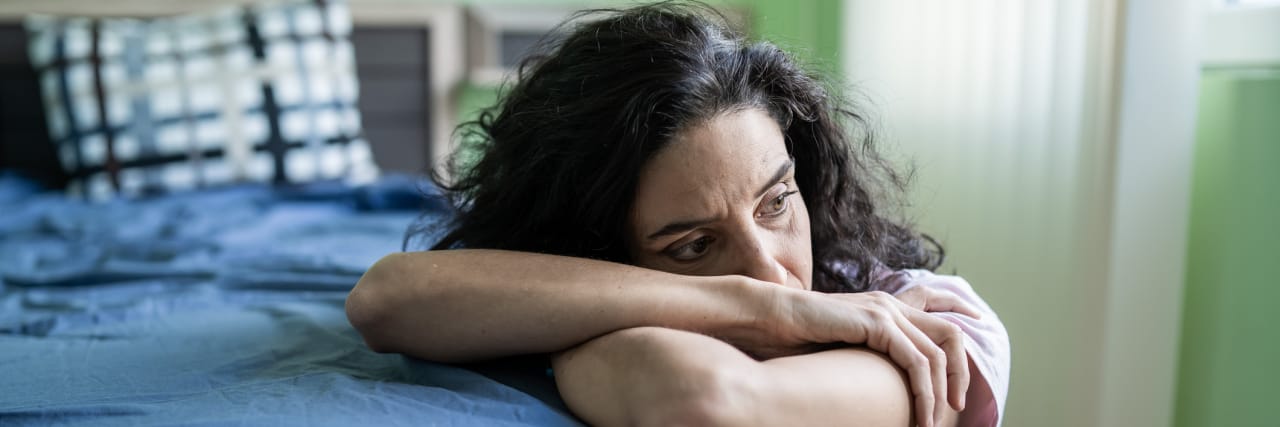 When Grief Doesn't Get Easier With Time Woman sitting next to a bed with arms folded, looking away with a sorrowful expression