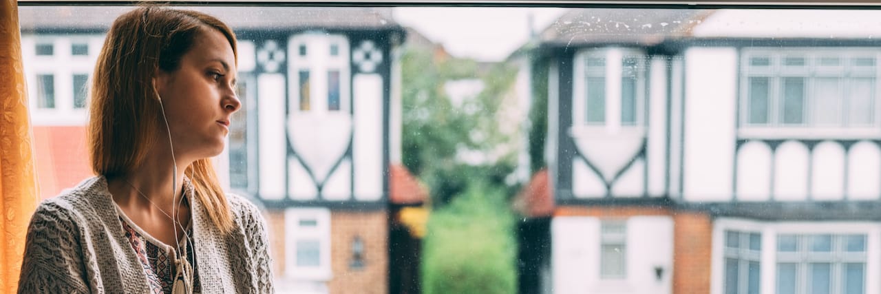 UK Mental Health Services Are Failing Those With Mental Illness Woman sitting in window with headphones on at home, in front of black and white Tudor buildings across the street