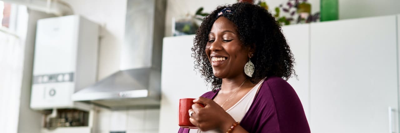 Black woman smiling, drinking coffee in kitchen and looking down at computer