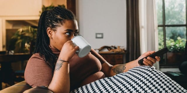 Dark skin woman is watching TV and drinking a coffee on the sofa