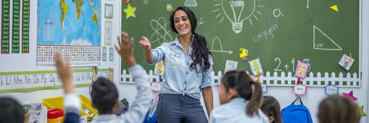 A woman of color stands at the front of an elementary classroom