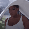 Megan Thee Stallion staring at the camera in all white at a funeral with a big floppy hat on. It's raining and everyone is wearing Black behind her.