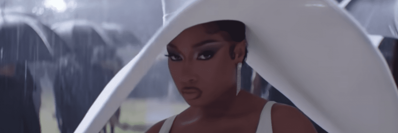 Megan Thee Stallion staring at the camera in all white at a funeral with a big floppy hat on. It's raining and everyone is wearing Black behind her.