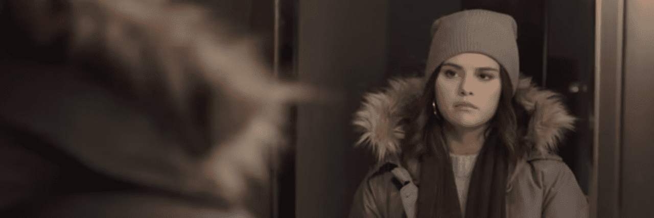 Selena Gomez in a winter jacket and beanie looking at herself in the mirror, serious expression