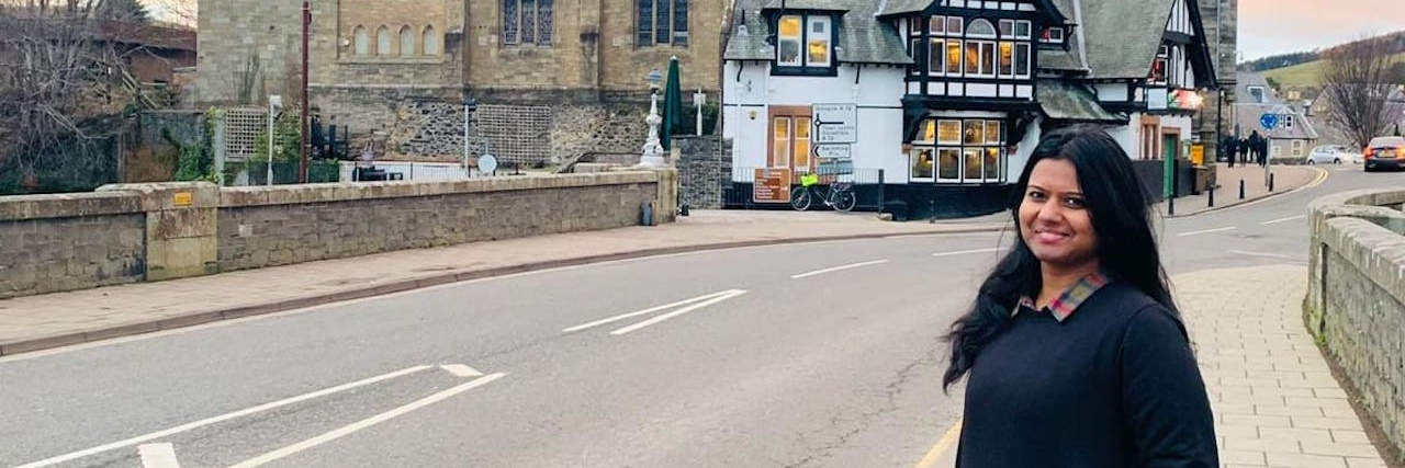 How Writing for Mental Health Became My Biggest Coping Mechanism Photo of contributor, an Indian woman, standing in front of bridge and black and white Tudor building in England