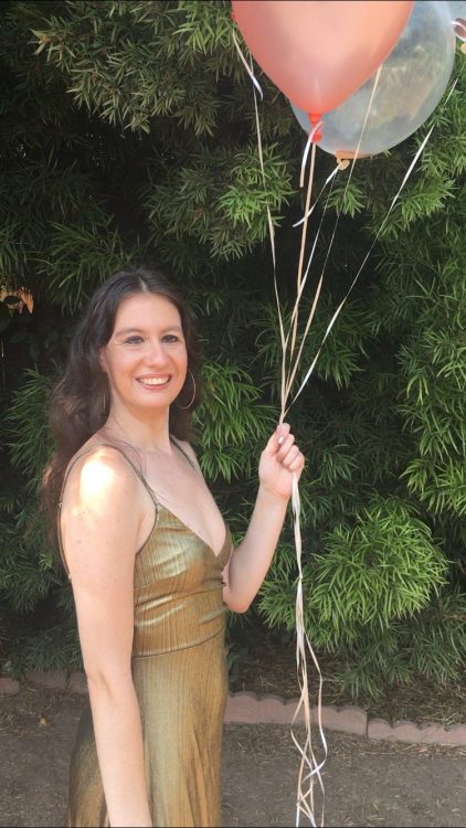 The author stands outside wearing a gold dress with spaghetti straps and holding pink and white balloons.
