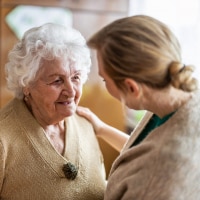 A woman with blonde hair speaks to an elderly woman wearing a beige top.