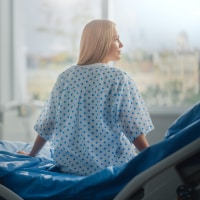 A young woman with blonde hair looks out a window while sitting on a hospital bed and wearing a hospital gown.