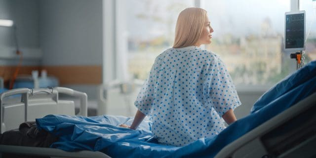 A young woman with blonde hair looks out a window while sitting on a hospital bed and wearing a hospital gown.
