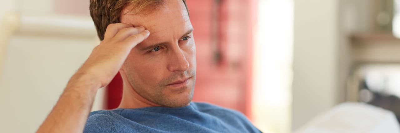 Reminders for Coping With Your Grief or a Loved One's Grieving Process A man with brown hair wearing a bliue shirt sits on a couch and stares pensively ahead with his hand on his forehead.
