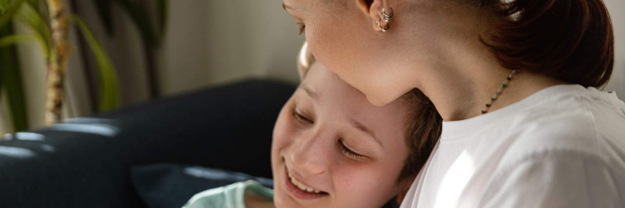 A woman with brown hair wearing a white top holds an orange inhaler and puts her arm lovingly around a boy with light brown hair.