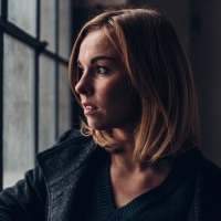 A woman with red hair wearing a dark coat looks out a window.