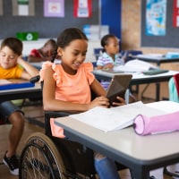 A black girl wearing an orange shirt and jeans sits in a wheelchair in a full classroom.