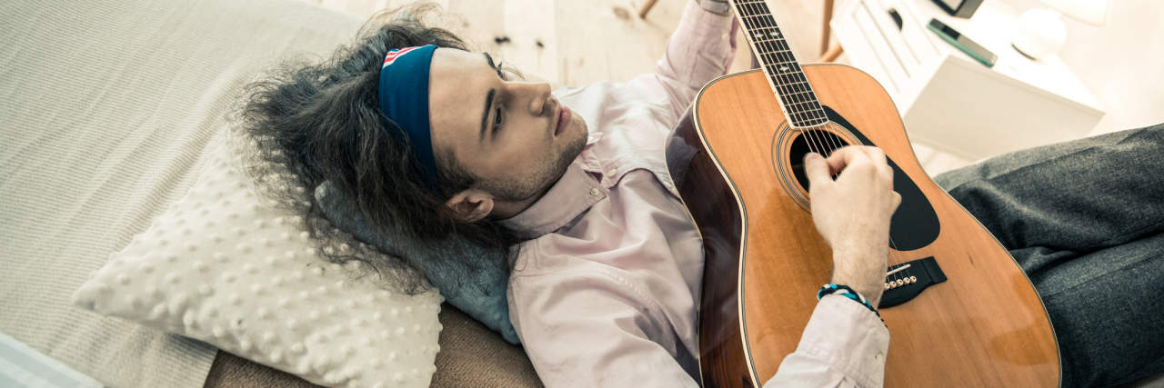 Young man with long curly hair lying on the ground playing guitar, sad