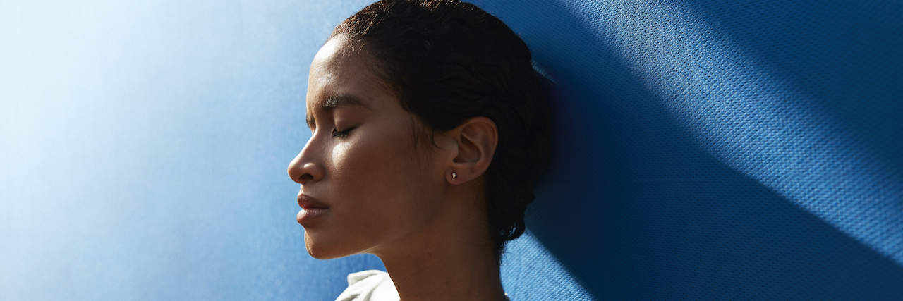 What the Media Always Gets Wrong About Chronic Illness Woman of color standing against blue wall with eyes closed