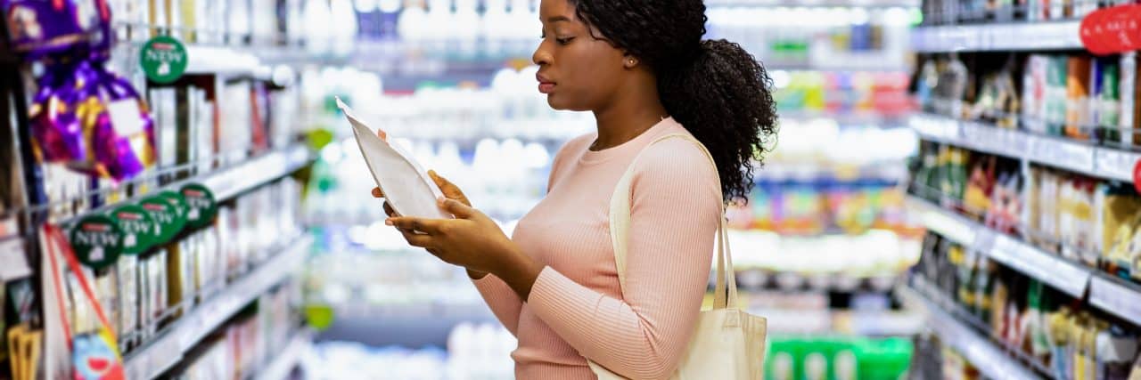 Pretty black lady with eco bag reading labels on food products, shopping for groceries at mall