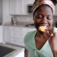 Close-up of mid adult black woman standing in kitchen eating a cracker with a slice of Gouda cheese