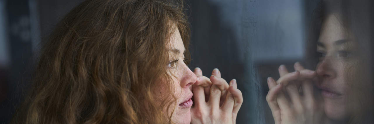 Woman with hands clasped, looking out of window and her reflection in it