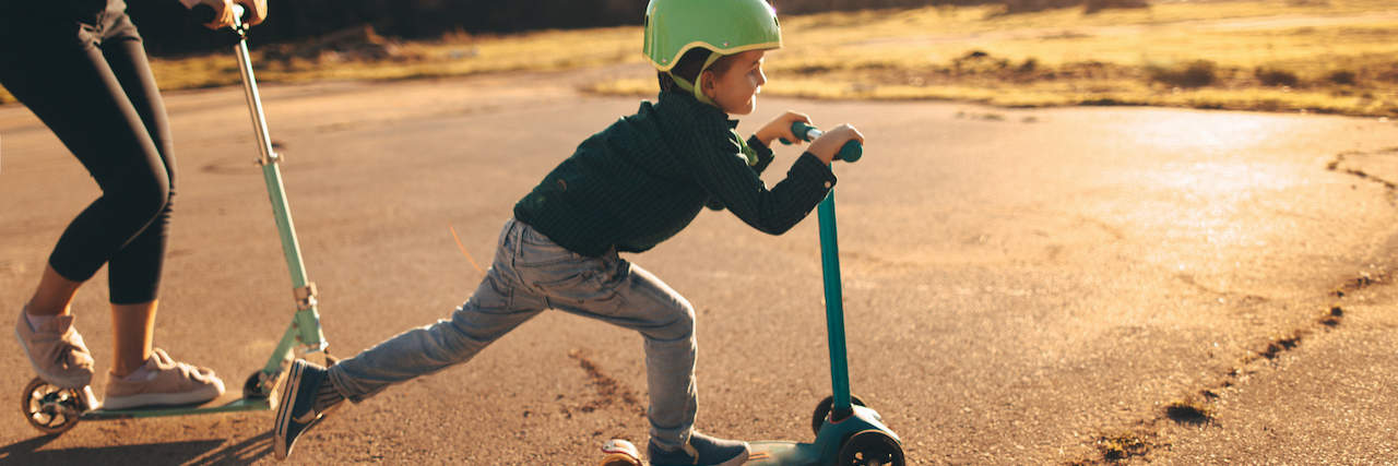 Young boy riding a push scooter, with his mom on a scooter behind him