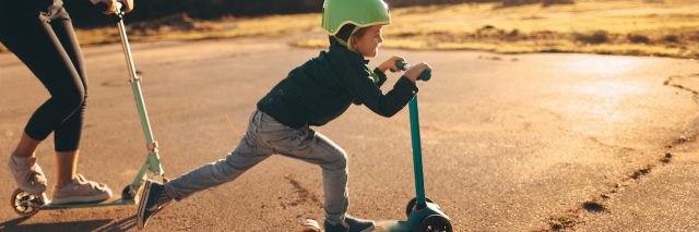 Young boy riding a push scooter, with his mom on a scooter behind him