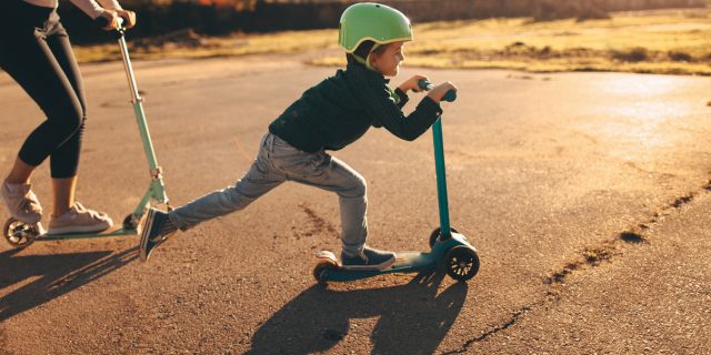 Young boy riding a push scooter, with his mom on a scooter behind him