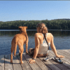 A golden yellow pit bull/vizsla mix and a girl with a ponytail sitting on the edge of a dock, looking out over a lake