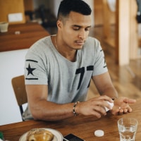 A man of color wearing a gray sports jersey pours pills into his hand while sitting at a table.