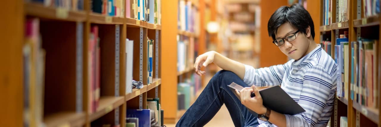 Studying Philosophy to Help Mental Health With Schizoaffective Disorder A man sits between rows of bookshelves while reading a book.