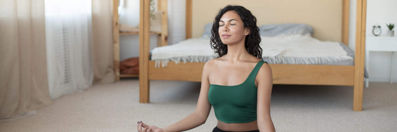 A woman with curly brown hair wearing a green top and black yoga pants sits in front of a bed in a meditative pose.