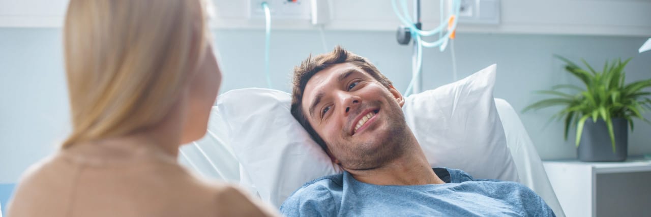 Learning to Ask for Help in Life With Functional Neurological Disorder and Chronic Pain A man with brown hair lies in a hospital bed while a woman with blonde hair holds his hand.