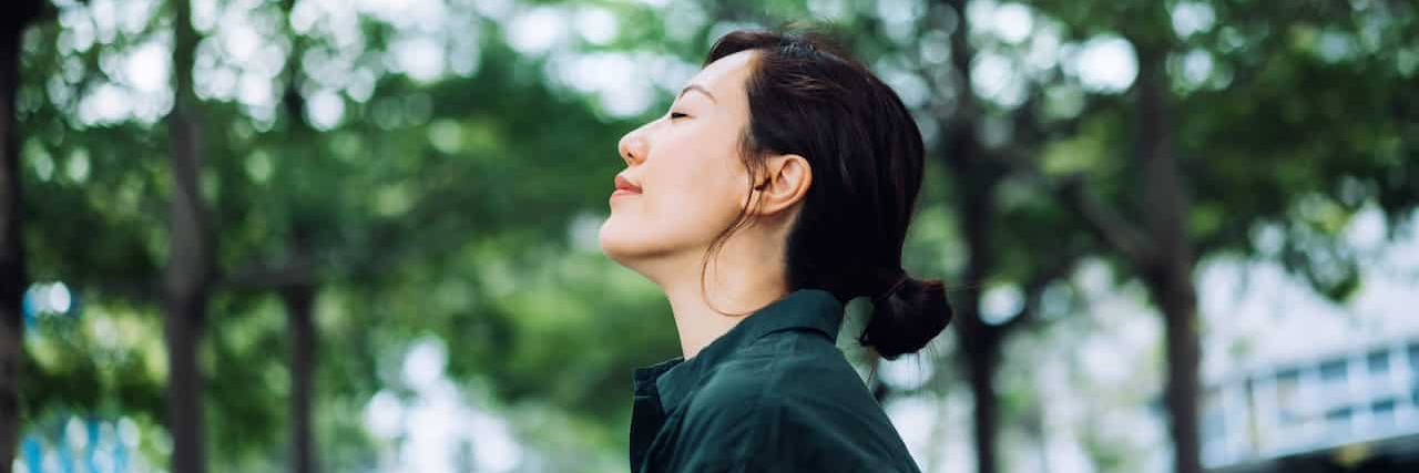 Asian woman standing outside in front of trees with her eyes closed, looking peaceful