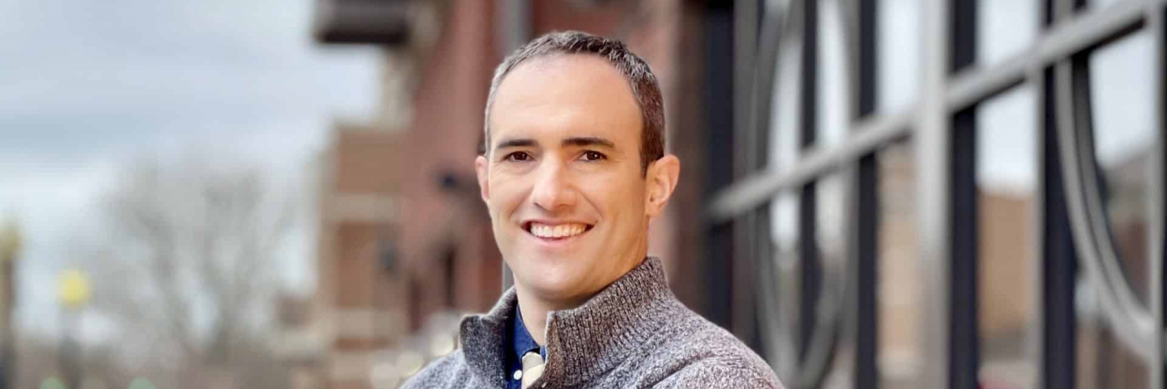 Challenging the Perception that Psychiatrists are 'A Little Robotic' Dark-haired man wearing sweater stands in front of building with tall glass windows