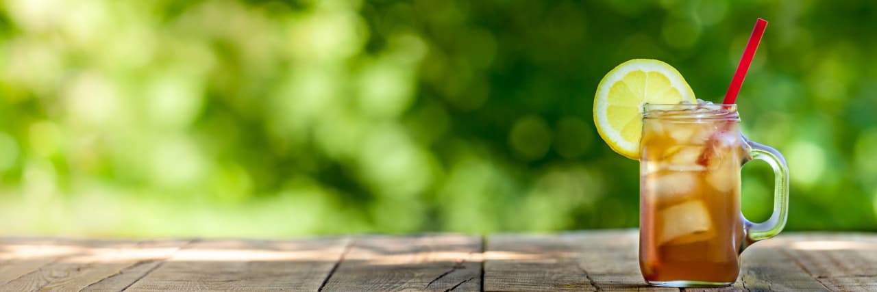 When Life Gives You Lemons, Make Sweet Tea: How I Survived My Brother's Brain Tumor glass of iced tea with lemon wedge on rim sitting on wooden table in front of green leafy background.