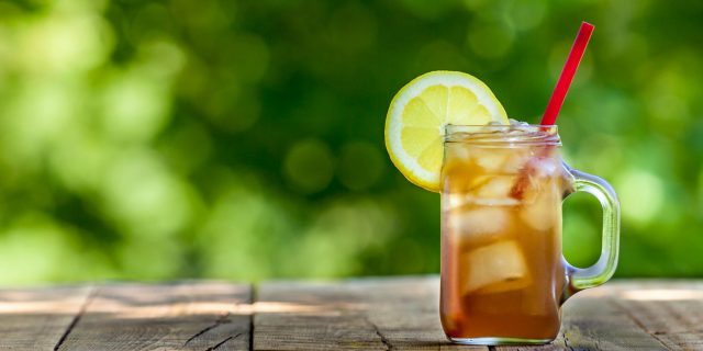 glass of iced tea with lemon wedge on rim sitting on wooden table in front of green leafy background.