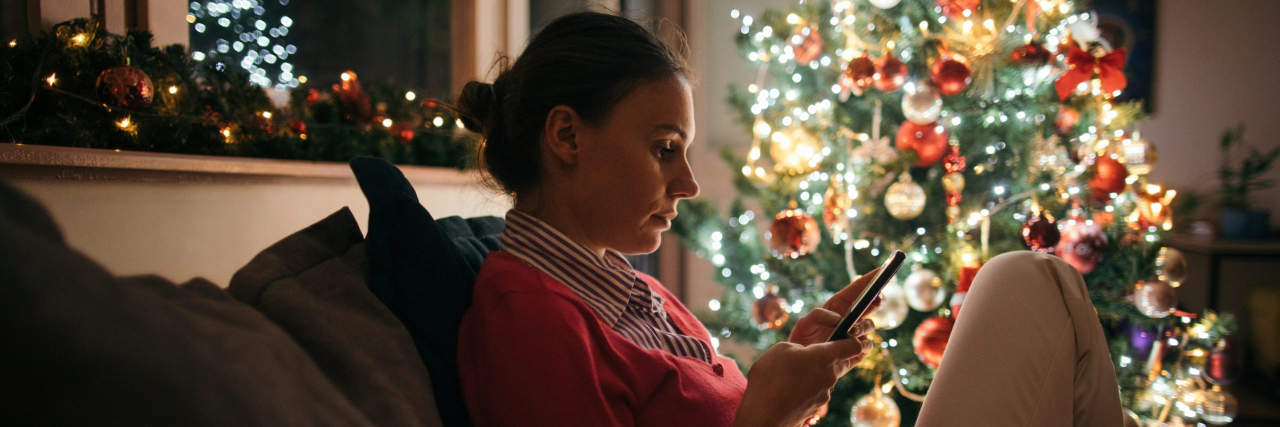 Young woman sitting alone on the sofa next to a decorated Christmas tree and using mobile phone