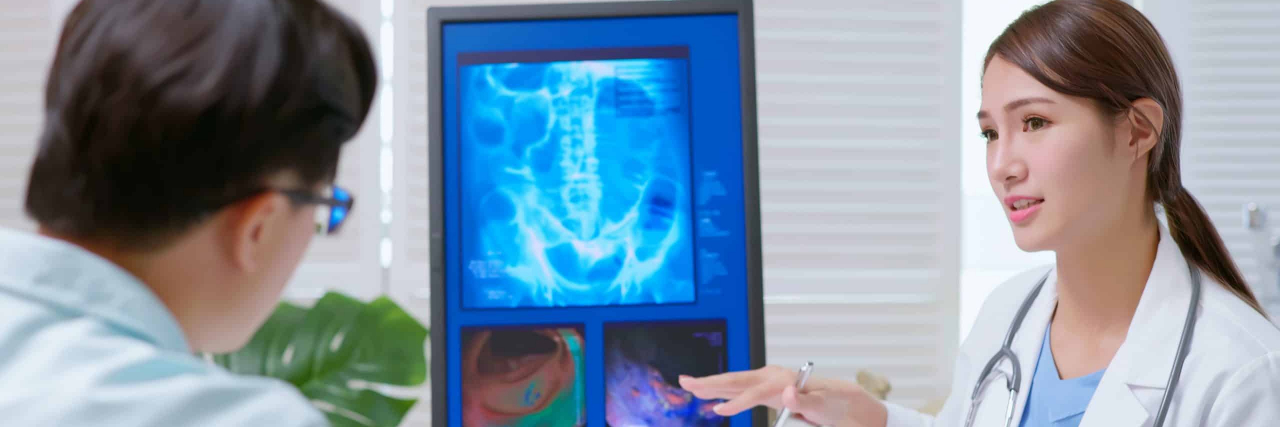 A Beginner's Guide to Your First Colonoscopy young brunette ponytail female asian doctor wear white coat and stethoscope pen pointing explaining xray film on computer to male patient at clinic - an colon model in background