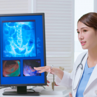 young brunette ponytail female asian doctor wear white coat and stethoscope pen pointing explaining xray film on computer to male patient at clinic - an colon model in background